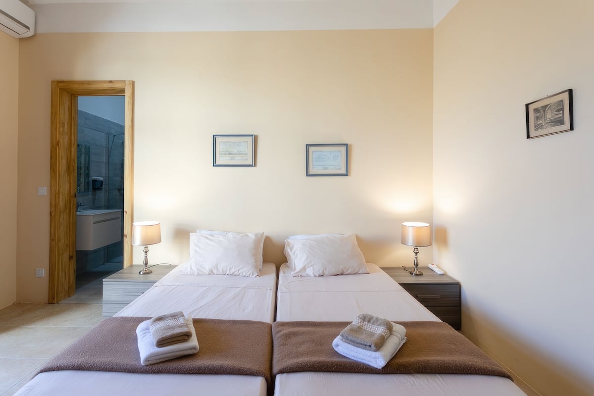 A hotel room with two beds, each with a pillow and a blanket, and two bedside lamps. There are framed pictures on the wall and a wooden door leading to a bathroom.