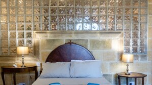 A neatly made bed with two pillows, a blue blanket, and two folded towels. There are two bedside tables with lamps and a phone on one. The wall behind the bed has a decorative glass panel. - Villa Serenity B&b (Sannat)