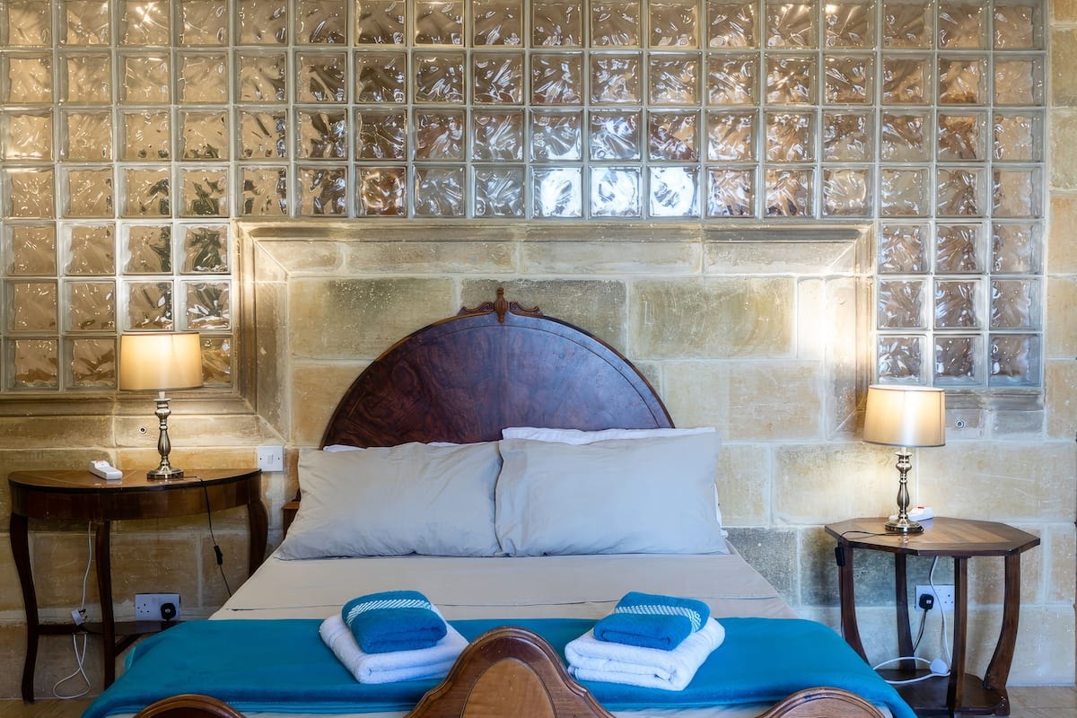 A neatly made bed with two pillows, a blue blanket, and two folded towels. There are two bedside tables with lamps and a phone on one. The wall behind the bed has a decorative glass panel.