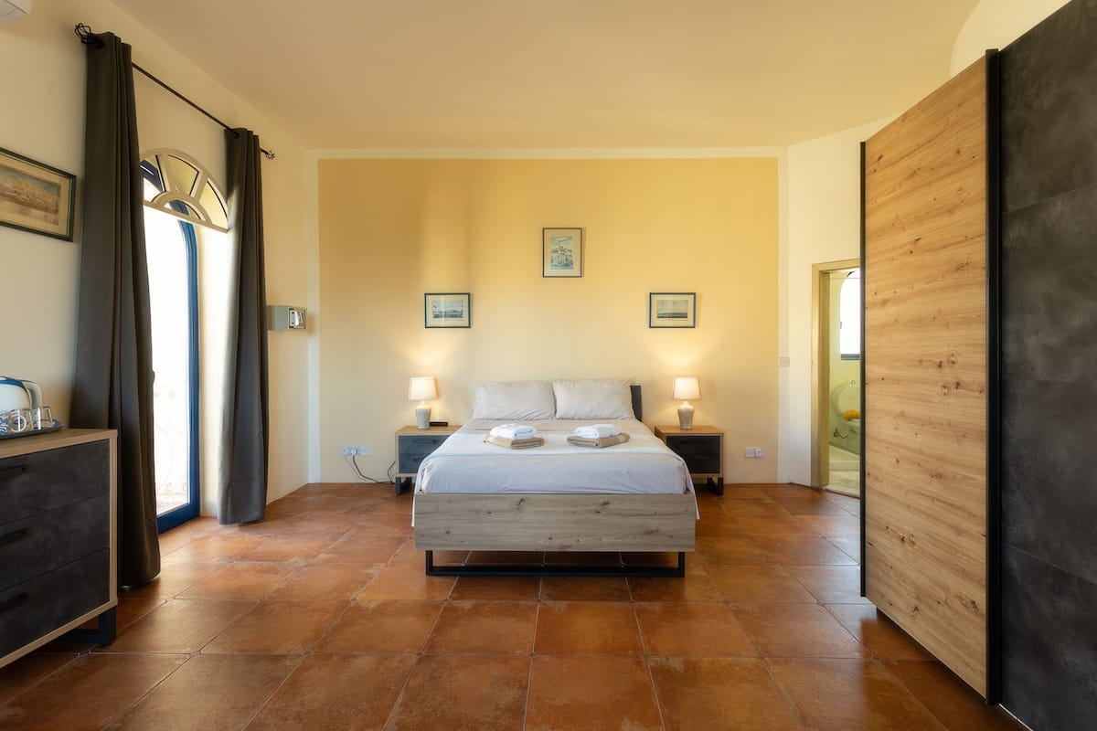 A bedroom with a large bed, two bedside tables with lamps, a wooden wardrobe, and a door leading to another room.