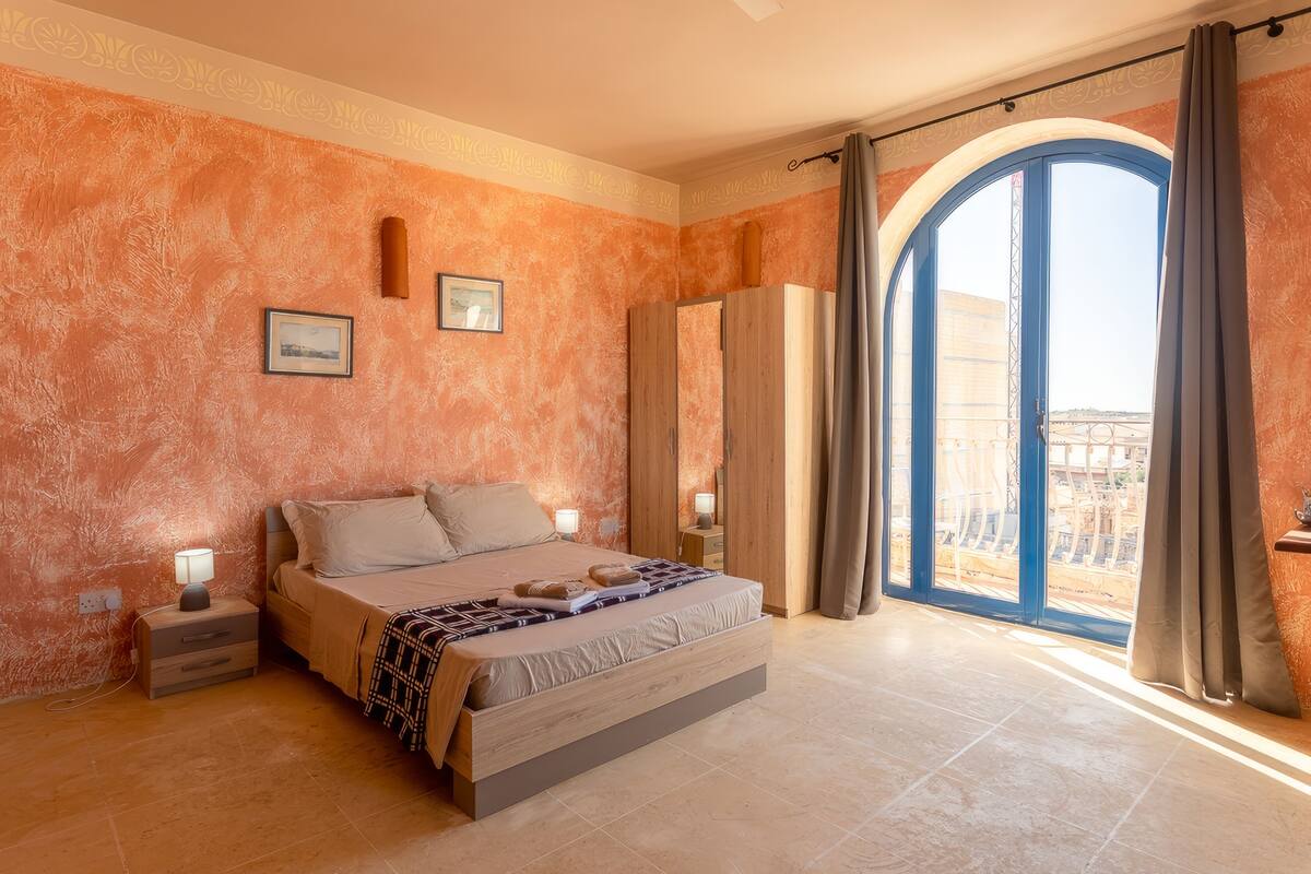 A bedroom with a large bed, a nightstand, a wardrobe, and a view through a large window.