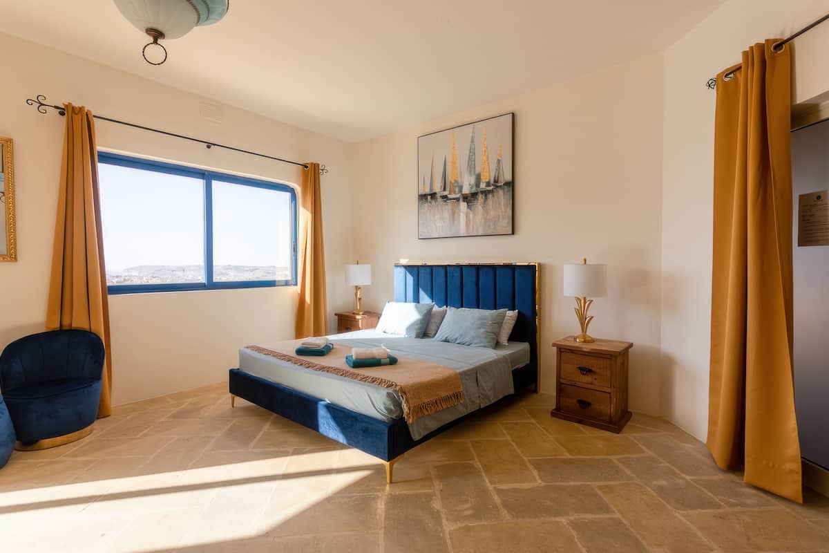 A bedroom with a large bed, a chair, a bedside table, and a window with curtains.