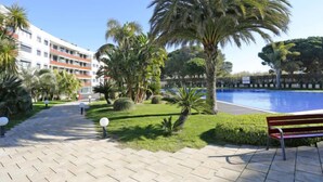 Zona junto a la piscina con un banco, palmeras y un edificio al fondo.