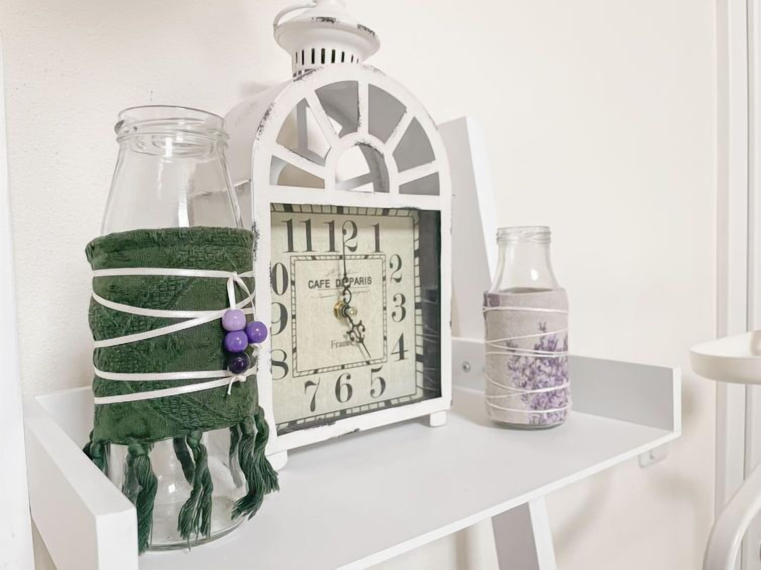 A white table with a clock, a jar with a green cloth, and a bottle with purple flowers.