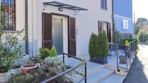 Un immeuble résidentiel doté d’une porte d’entrée, de deux fenêtres avec volets, d’un petit espace extérieur aménagé avec une table et des chaises, et de plantes en pot.