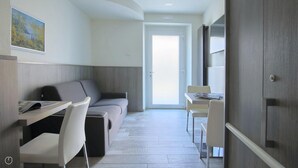 Une pièce moderne et bien éclairée, comprenant un canapé, un bureau et une chaise, et dotée d’une grande fenêtre et d’une porte donnant sur l’extérieur.