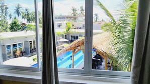 Vista a través de una ventana de un resort con piscina, palmeras y una estructura de techo de paja.