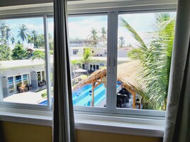 A view through a window of a resort with a pool, palm trees, and a thatched-roof structure.