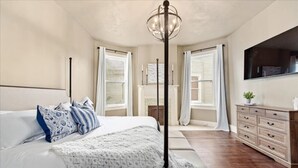 A bedroom with a large bed, a bedside table, a dresser, a TV mounted on the wall, and a window with curtains. - Downtown Heritage Suite | Free Parking (Macon)