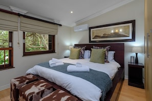 Une chambre d’hôtel avec un lit, une table de chevet, une lampe, un tableau encadré et une fenêtre donnant sur un paysage verdoyant.
