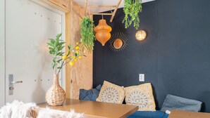 A cozy room with a wooden table, a blue sofa with patterned cushions, a wicker vase with yellow flowers, and hanging plants.