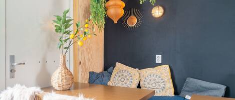 A cozy room with a wooden table, a blue sofa with patterned cushions, a wicker vase with yellow flowers, and hanging plants.