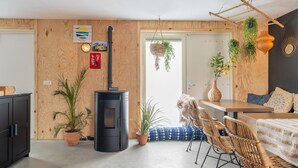 A cozy living room with a wood-paneled wall, a black wood-burning stove, wicker furniture, and hanging plants.