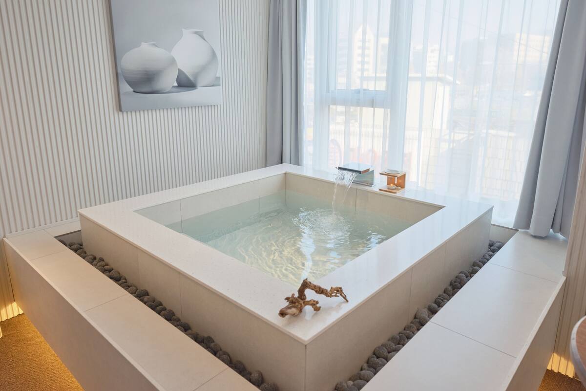 Un baño moderno con una bañera grande, un lavamanos pequeño y un ventanal con cortinas.