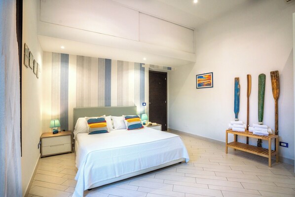 A modern bedroom with a large bed, striped curtains, a nightstand with a lamp, and a wooden shelf with towels.