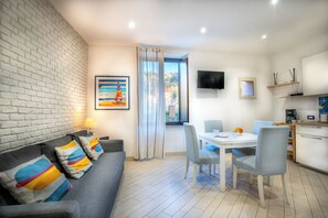 Ein modernes Wohnzimmer mit einem grauen Sofa, bunten Kissen, einer Essecke mit weißem Tisch und Stühlen, einem an der Wand befestigten Fernseher und einer Küchenzeile mit Schränken und einer Kaffeemaschine.
