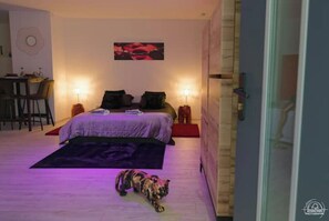 A bedroom with a bed, a desk, and a chair. There is a painting on the wall and a dog on the floor.