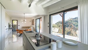 Een moderne keuken met een groot eiland, een eethoek met oranje stoelen, en een uitzicht op een glooiend landschap door grote ramen.