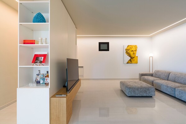 A modern living room with a grey sectional sofa, a flat-screen TV on a wooden stand, a bookshelf with various items, and a framed artwork on the wall.