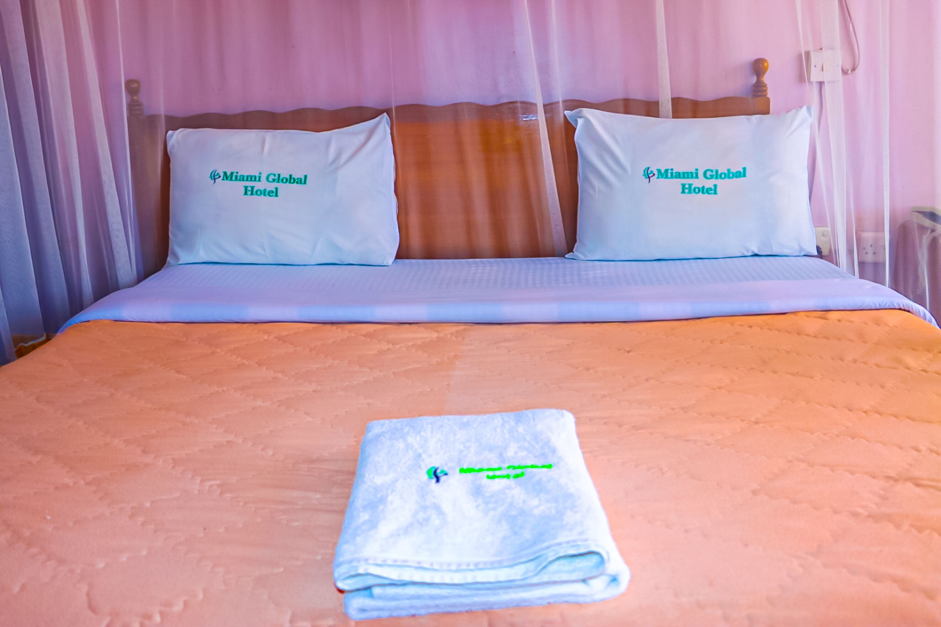 A neatly made bed with a folded towel and two pillows labeled "Miami Global Hotel."