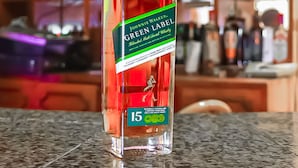 A bottle of Johnnie Walker Green Label whiskey on a countertop with a blurred background of shelves filled with various bottles. - Miami Global Hotel (Nairobi)
