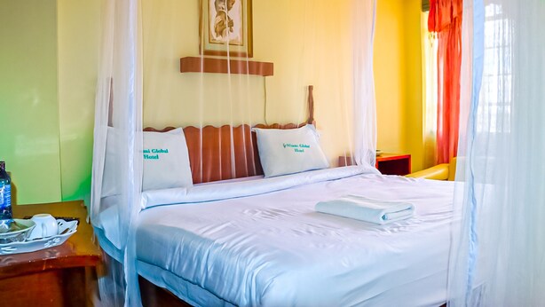 A hotel room with a bed, a bedside table, a nightstand, a framed picture on the wall, and a window with curtains. - Miami Global Hotel (Nairobi)