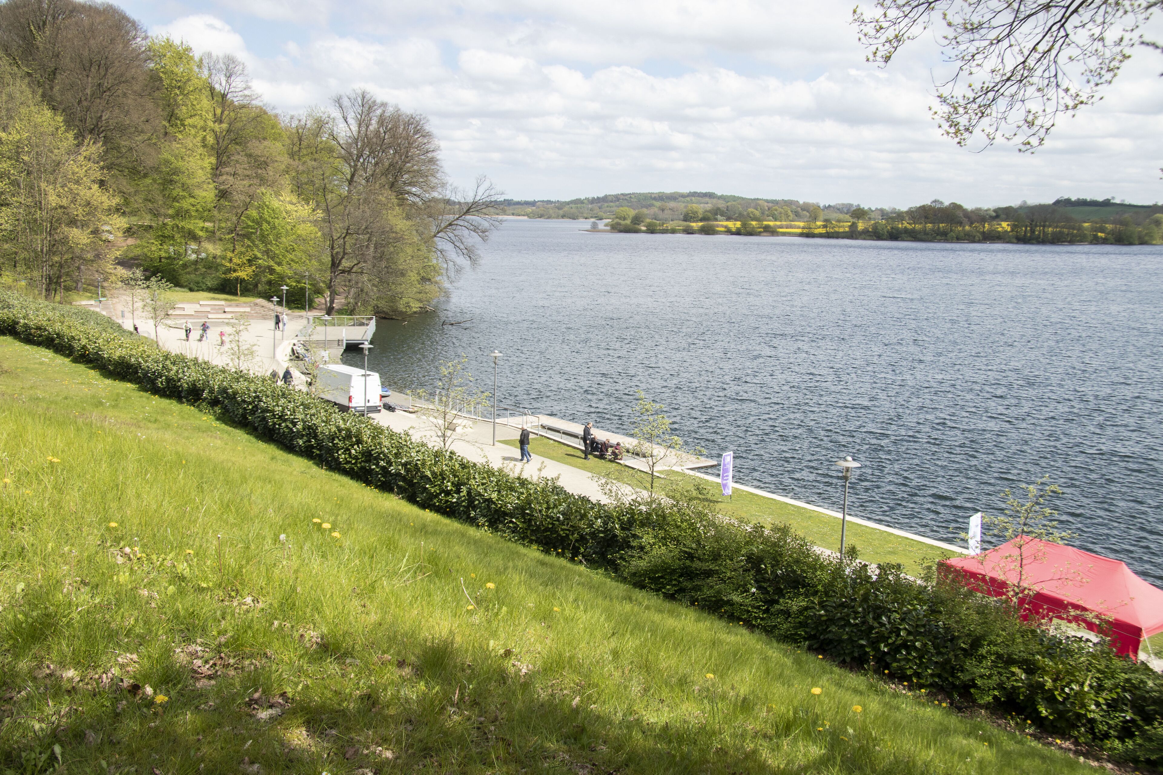 Ein Park am Flussufer mit einem Fußweg, Bänken und einem Gebäude mit rotem Dach.