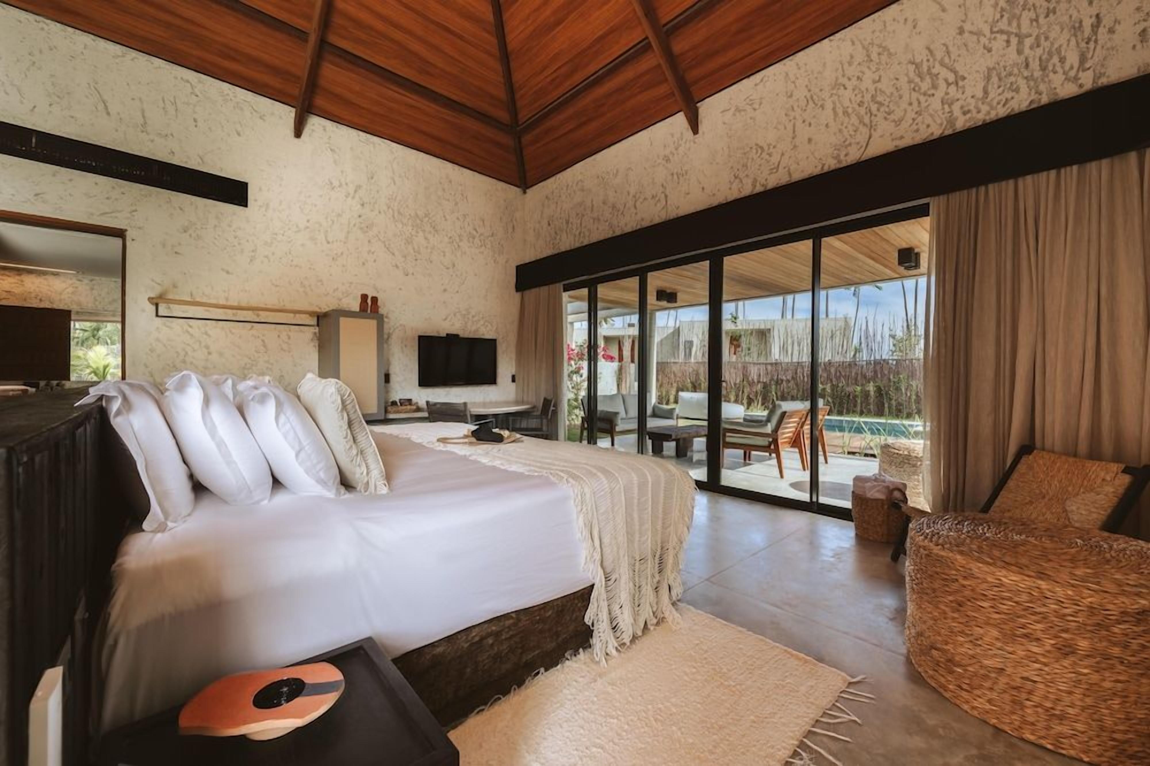 Quarto com uma cama grande, televisão e vista para área externa com assentos.