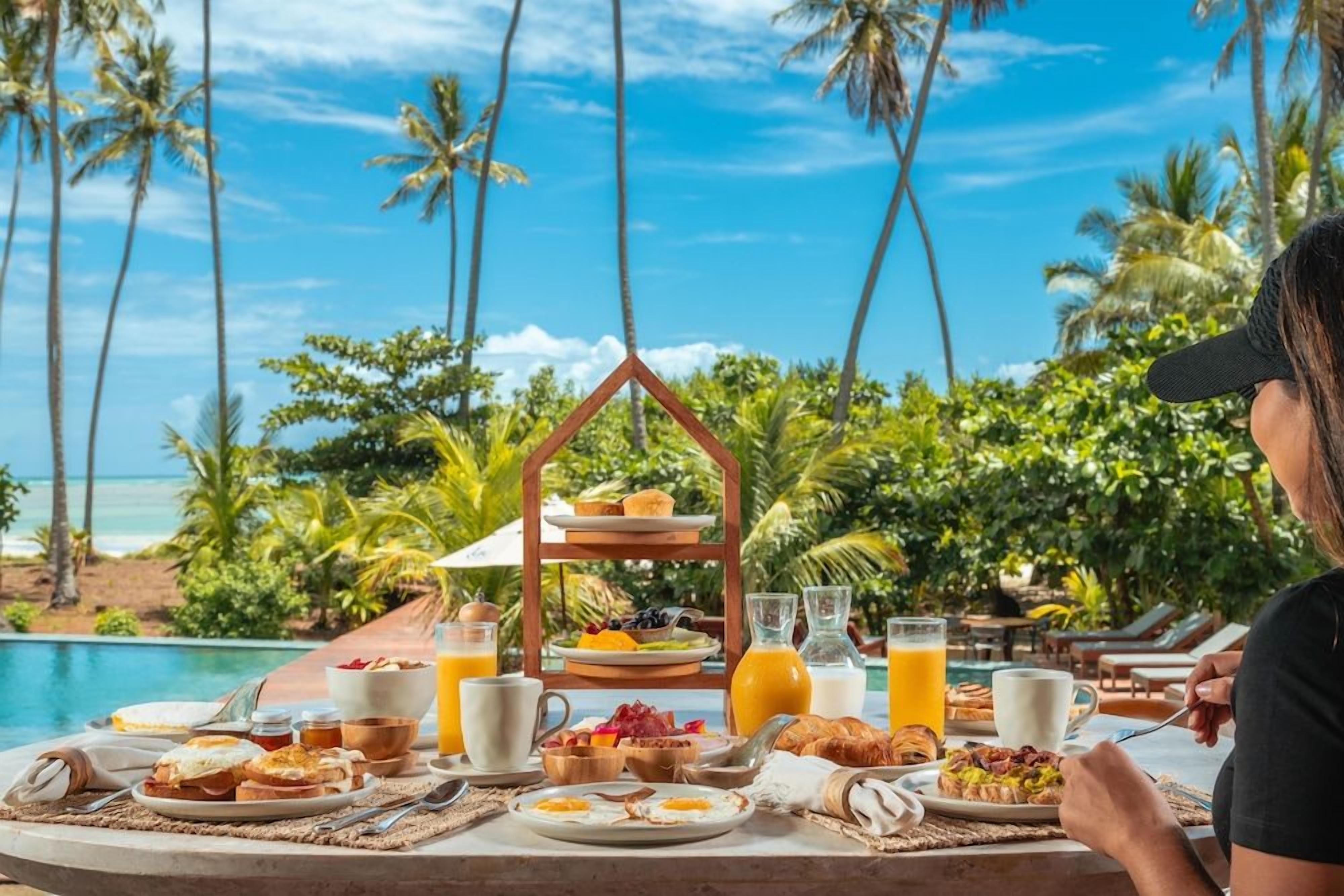 Mesa de café da manhã com variedade de alimentos e bebidas, um suporte em três níveis e vista para o oceano com palmeiras.