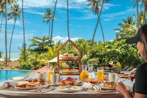 Ein Frühstückstisch reich an Speisen und Getränken, mit einem dreistufigen Etagere und Blick auf das Meer mit Palmen.