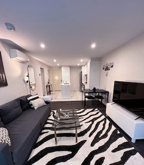 Een moderne woonkamer met een bank, een glazen salontafel, een vloerkleed met zebraprint, een televisie en een keuken op de achtergrond.