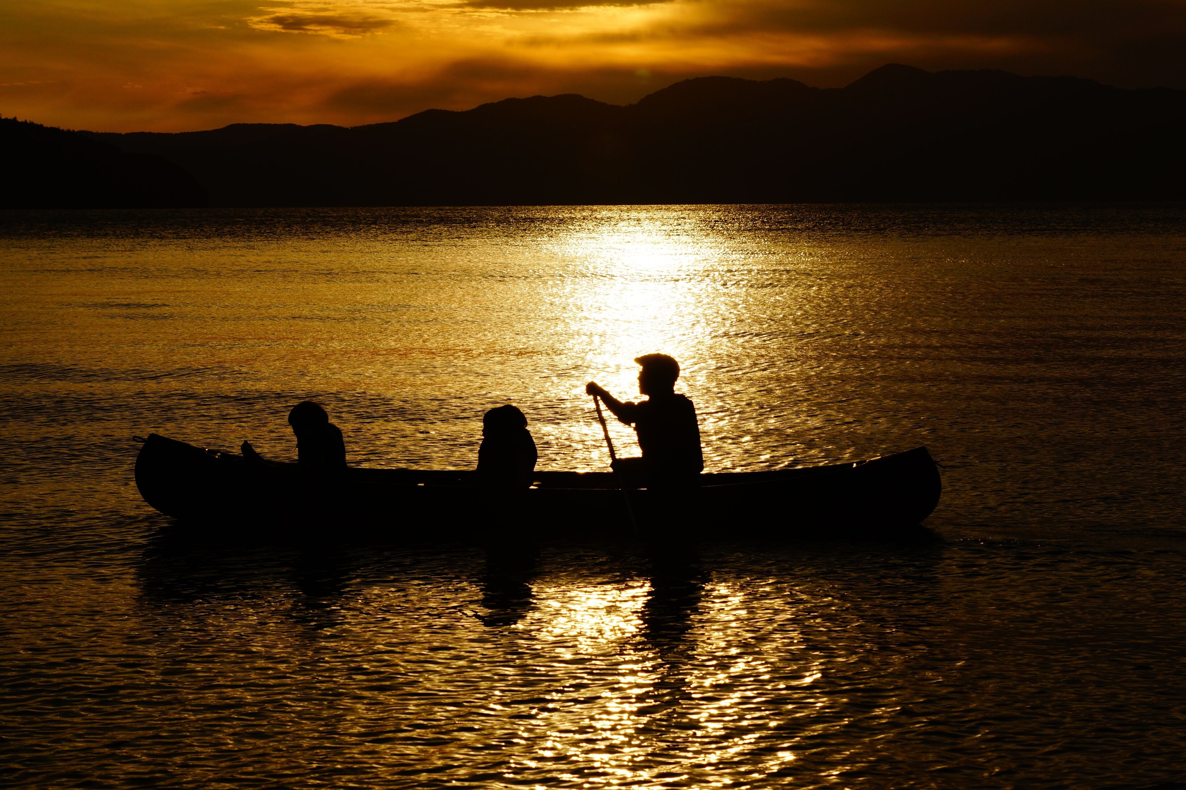 La silhouette de trois personnes dans un canoë, sur une étendue d’eau, au coucher du soleil.