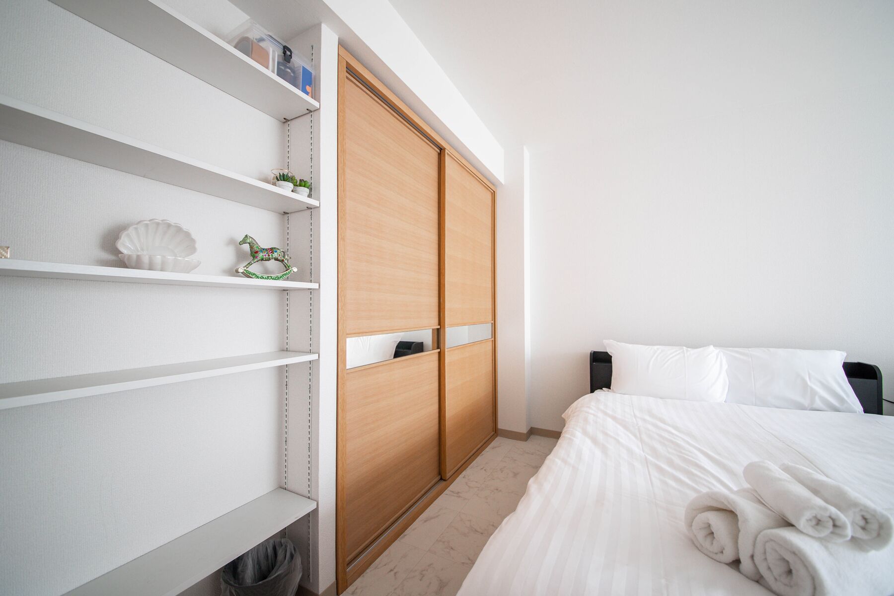 Een slaapkamer met een bed, witte beddengoed, een houten garderobe en planken met decoratieve objecten.