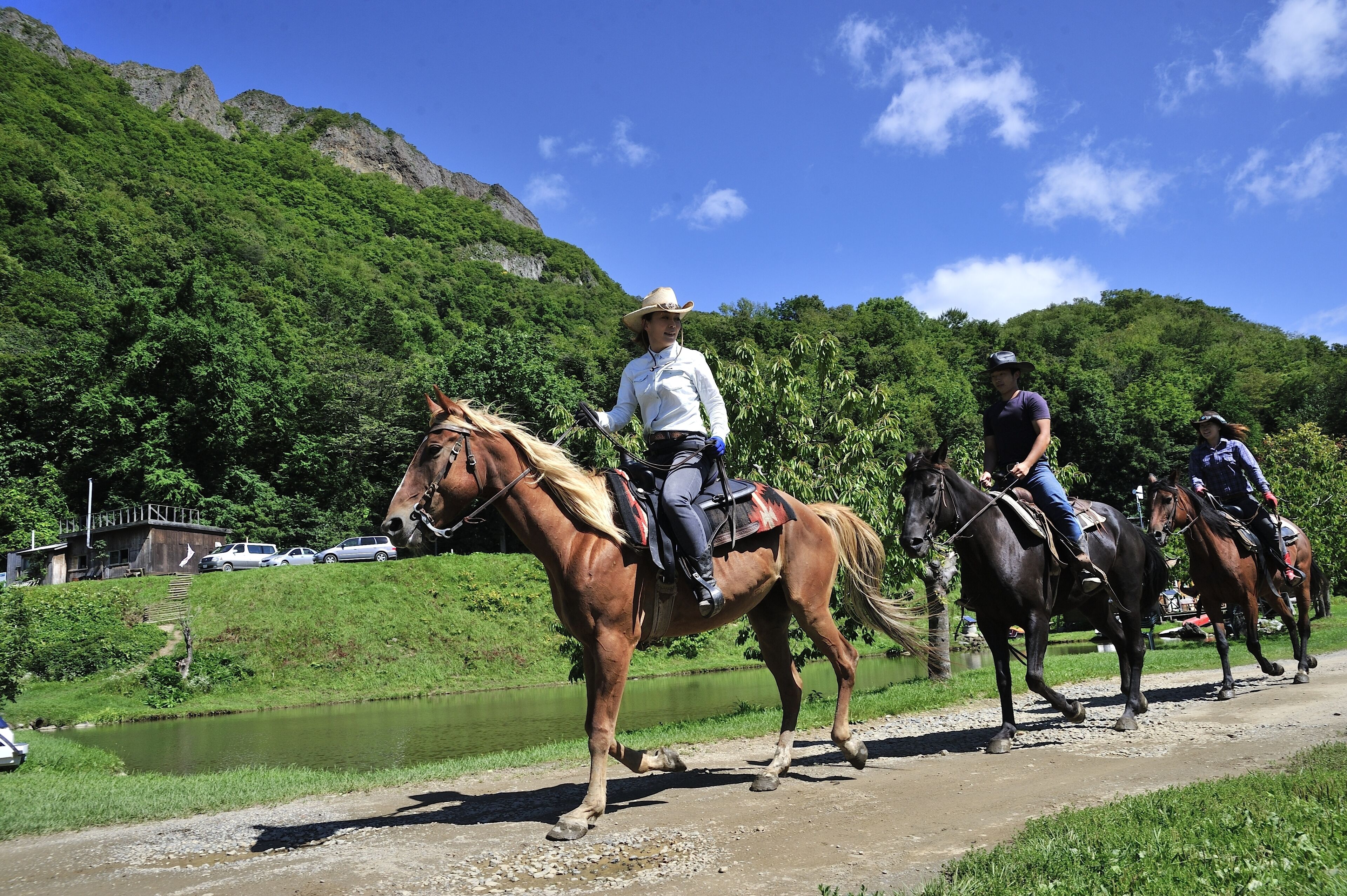 A group of people riding horses along a path with a river and green hills in the background.