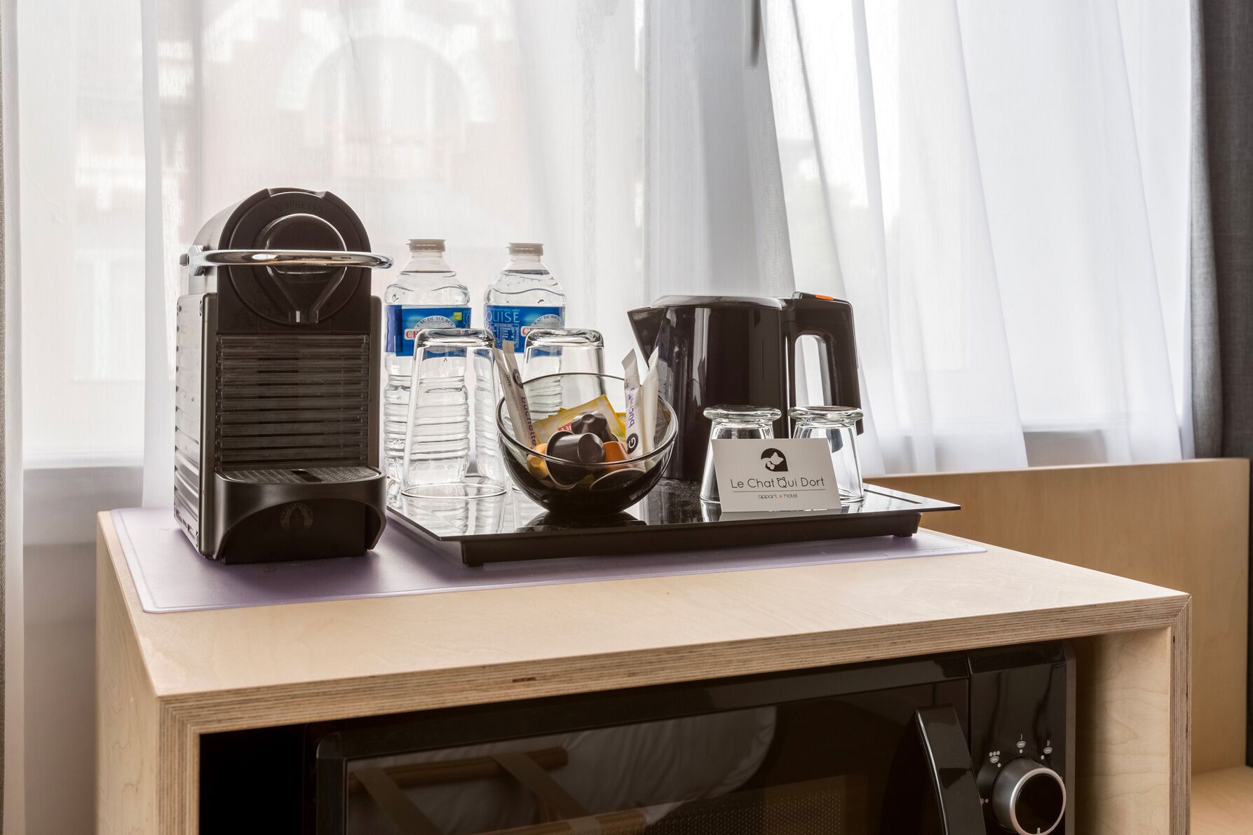 A coffee station with a coffee maker, water bottles, and a bowl of snacks on a wooden table.