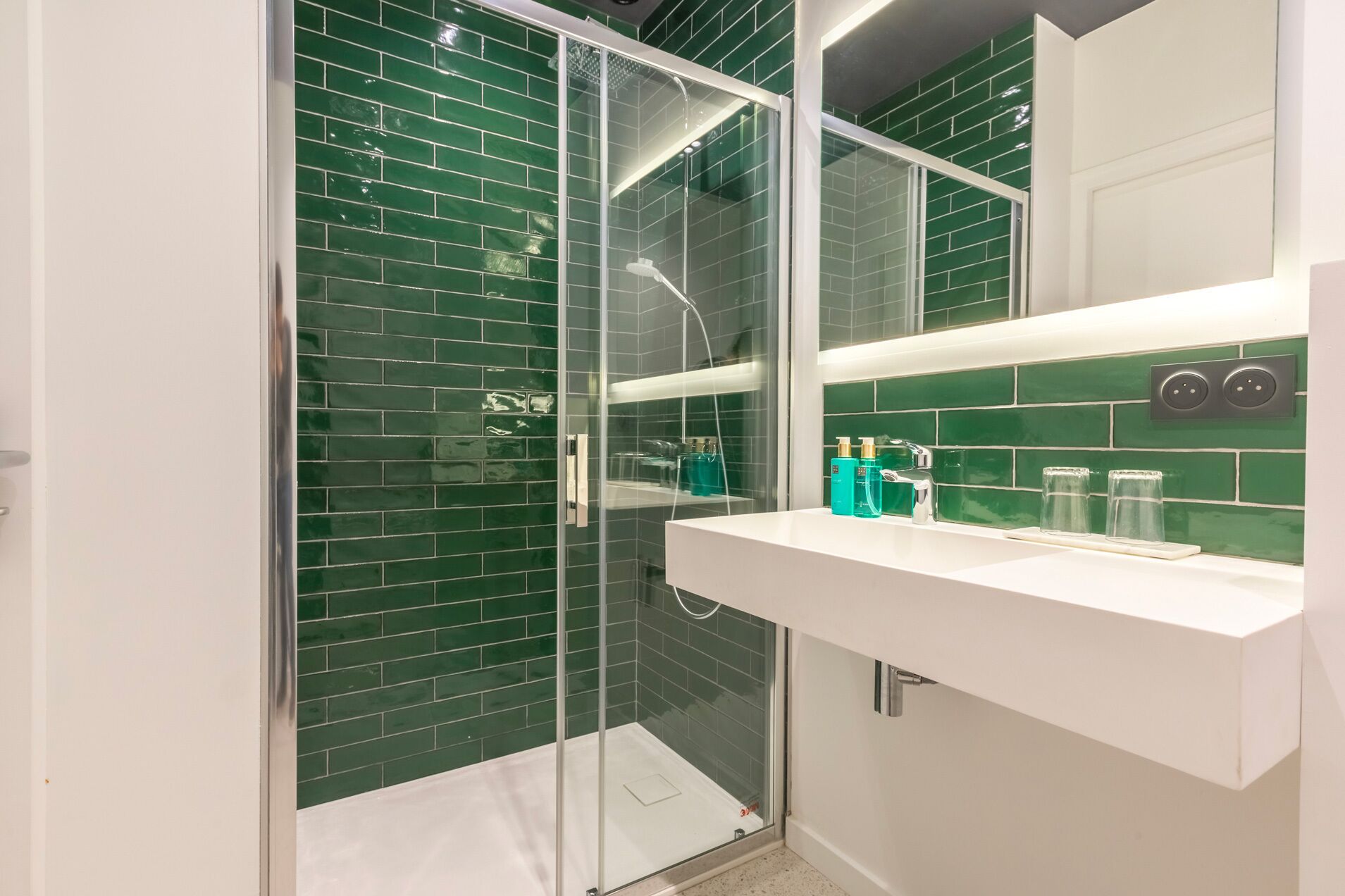 Un baño moderno con ducha envuelta en vidrio, paredes de azulejos verdes y un tocador blanco con artículos de aseo.