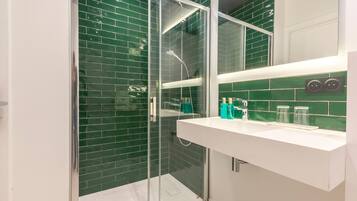 Un baño moderno con ducha envuelta en vidrio, paredes de azulejos verdes y un tocador blanco con artículos de aseo.