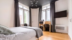 A modern hotel room with a large bed, a desk with a chair, a TV mounted on the wall, and a small kitchenette area.