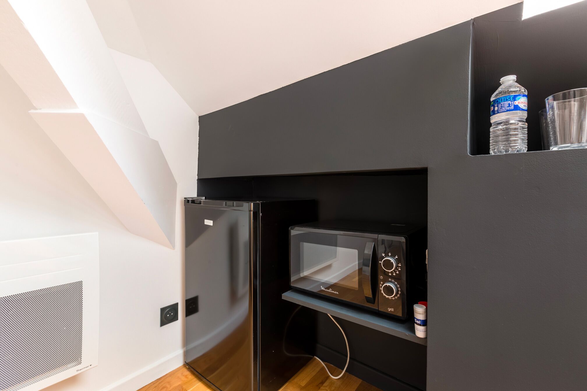 Una kitchenette compacta con refrigerador, microondas y una botella de agua sobre un estante.