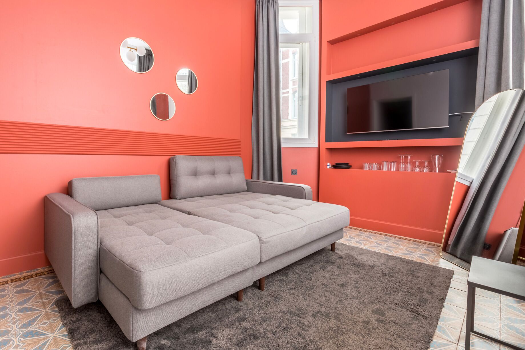A modern living room with a grey sofa, a flat-screen TV mounted on the wall, and a window with grey curtains.