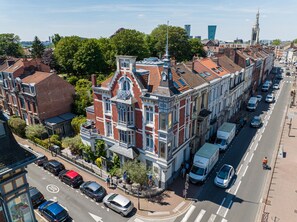Un edificio histórico con una torre destacada y ventanas amplias, situado a lo largo de una calle concurrida, donde se ven coches aparcados y un ciclista.
