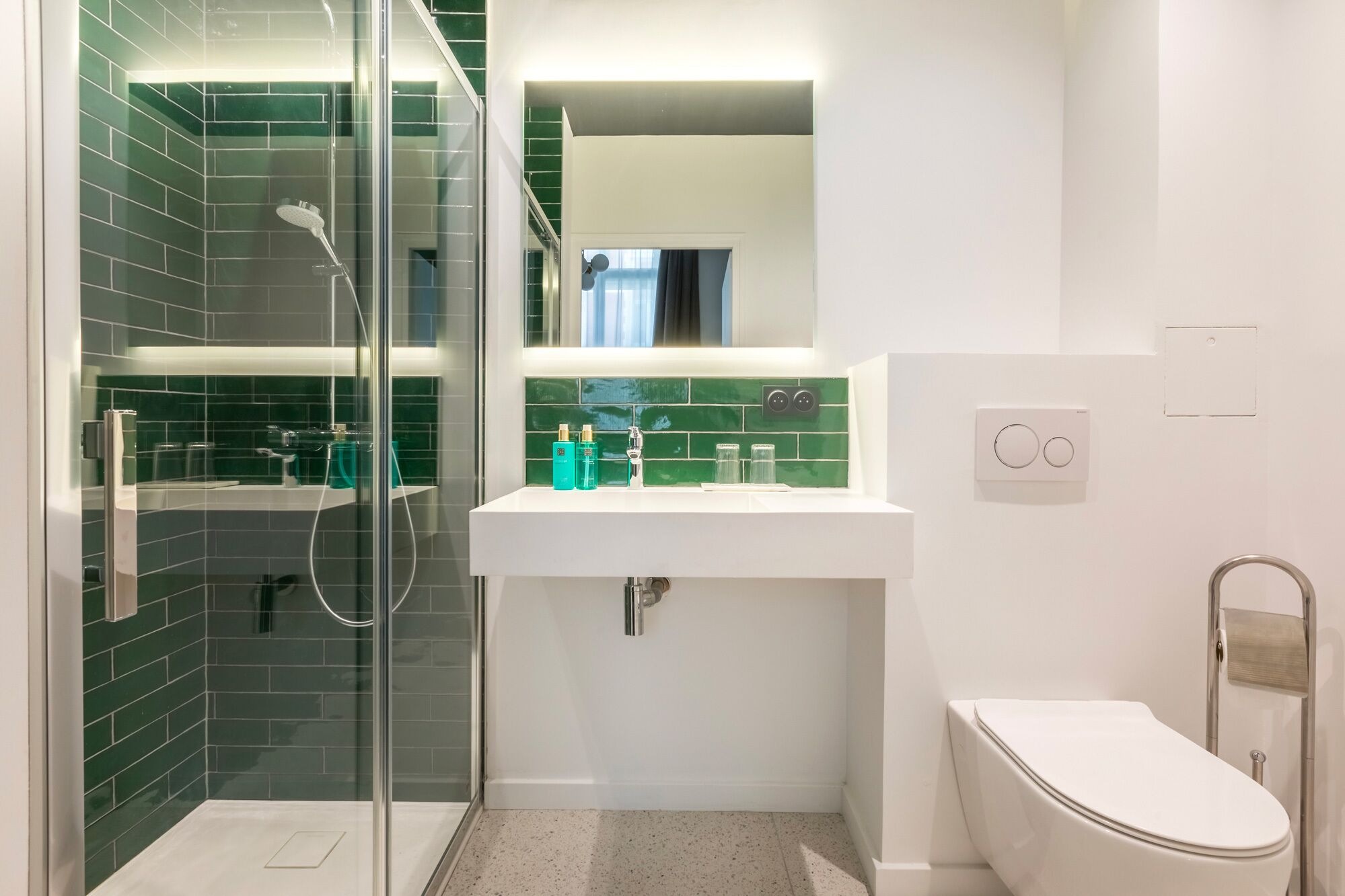 Casa de banho moderna com chuveiro separado por vidro, lavatório branco com azulejos verdes e sanita branca.