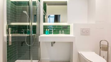 Casa de banho moderna com chuveiro separado por vidro, lavatório branco com azulejos verdes e sanita branca.