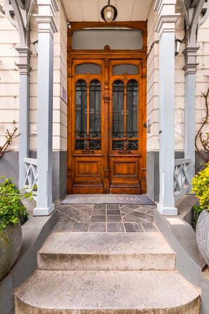 Una puerta doble de madera con paneles de vidrio, flanqueada por columnas blancas, que da acceso a una escalera de piedra.