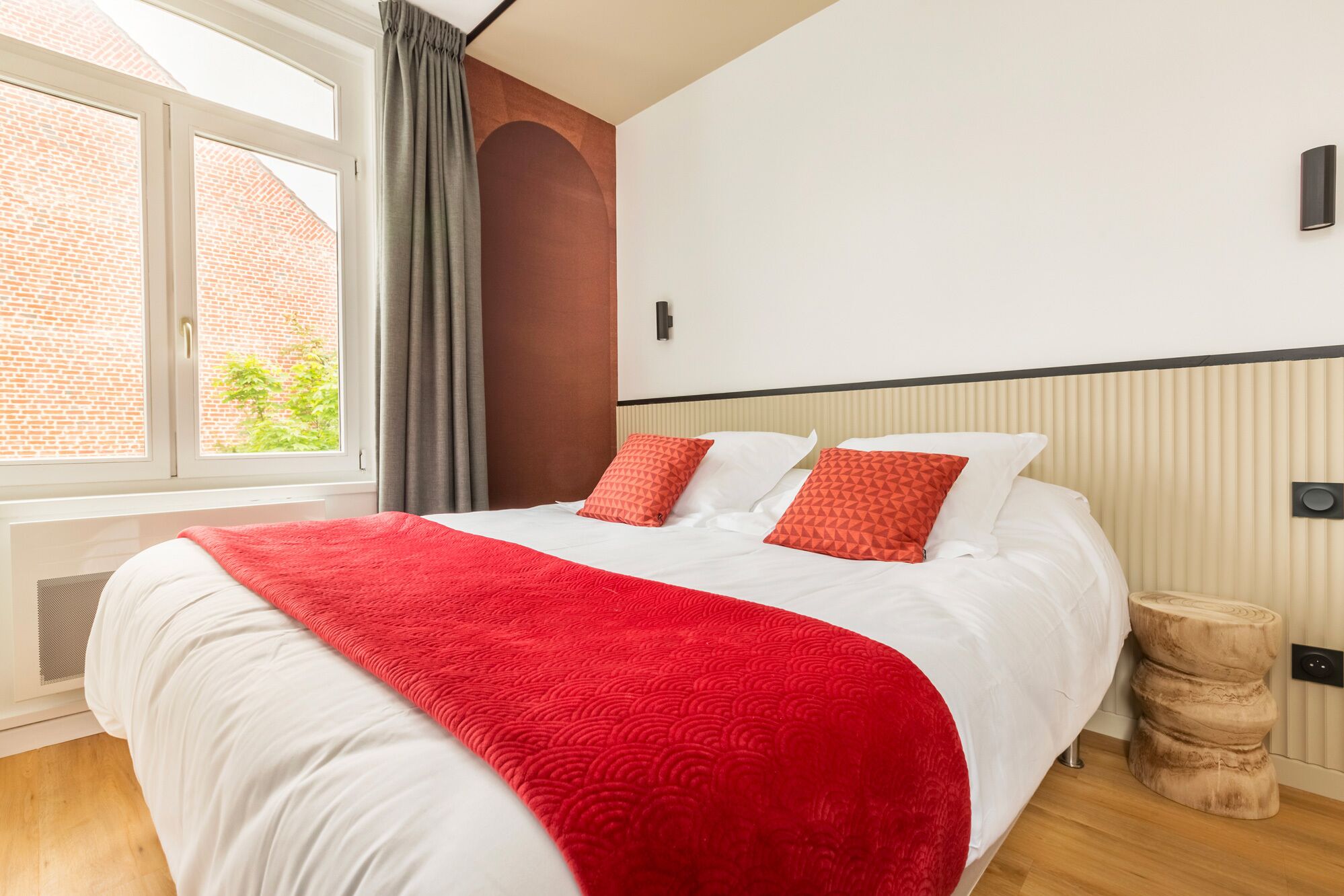 Une chambre d’hôtel moderne avec un grand lit, un plaid rouge et une vue sur un immeuble en briques par la fenêtre.