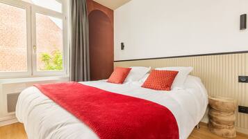 Une chambre d’hôtel moderne avec un grand lit, un plaid rouge et une vue sur un immeuble en briques par la fenêtre.
