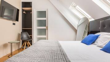 Un dormitorio con techo inclinado, una cama con almohadas azules, una silla, una mesita y un televisor en la pared.