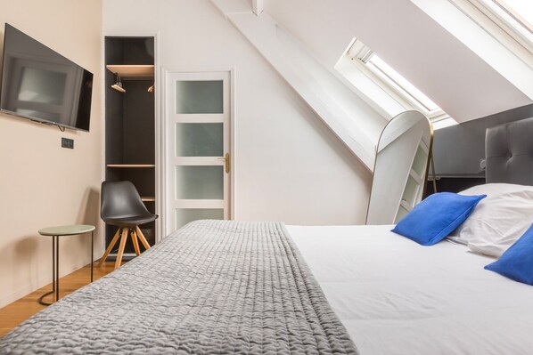 A bedroom with a sloped ceiling, a bed with blue pillows, a chair, a small table, and a TV on the wall. - Au Chat Qui Dort Lille - Villa Gounod (Lille)
