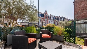 An outdoor patio with wicker furniture and a tiled floor, surrounded by greenery and a view of traditional buildings.