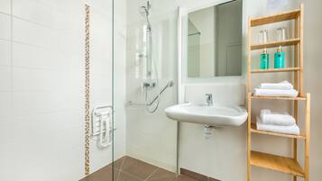 Un baño con ducha envuelta en vidrio, un lavamanos blanco, un espejo y un estante de madera con toallas.